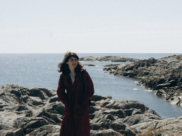 A woman in a long coat standing on a rocky seashore—alone in the open space, poised in a moment of transition