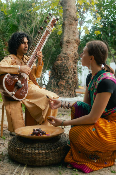 Dressed in traditional attire, an Indian musician plays the sitar outdoors during a festive gathering at home, while a woman in a saree lights a diya lamp to celebrate Diwali