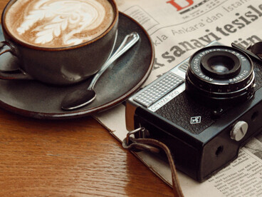 Un periódico y una cámara analógica acompañan una taza de café, evocando una época en la que el acceso a la actualidad estaba ligado a la lectura pausada y al trabajo paciente de quienes la registraban