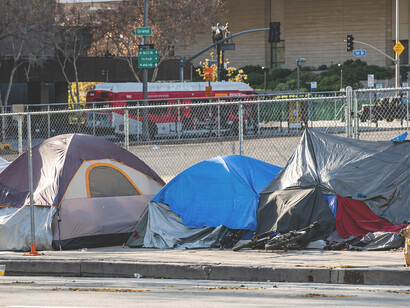 Homeless tents lined along the street beneath the shadow of modern luxury apartments, revealing the growing divide between wealth and housing insecurity