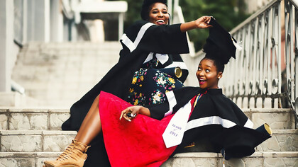Two women in academic attire pose in graduation robes on the stairs