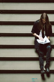 A person sitting on a set of stairs, holding a book, lost in thought or overwhelmed by the weight of their studies