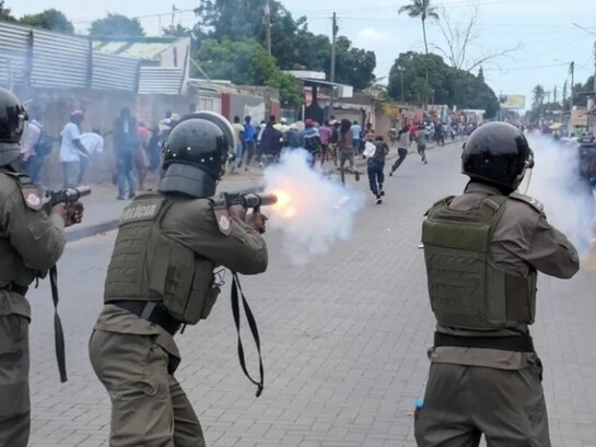 Policiais avançam e disparam contra manifestantes desarmados durante protestos de rua. A imagem traduz o clima de medo, repressão e instabilidade descrito no artigo, expondo a tensão política e social que tomou conta de Moçambique
