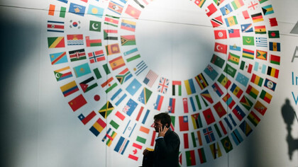 "A man in a black coat stands near a circular wall decorated in white, red, and blue, in front of a display of world flags
