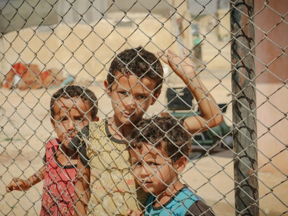Behind a chain lock fence in a Syrian refugee camp, children gather, their resilient spirits shining through despite the harsh realities they face