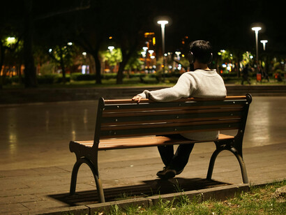 A solitary figure seated on a bench after dark in an urban park reflects the mood of isolation, the weight of urban loneliness, and the sense of living in a disconnected world