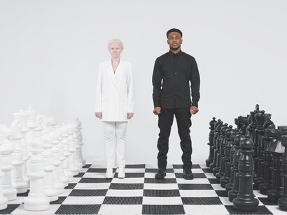 Standing on a sprawling chessboard that covers the floor, a woman in an immaculate white suit and a man in a sleek black suit exude an air of elegance and strategic intent