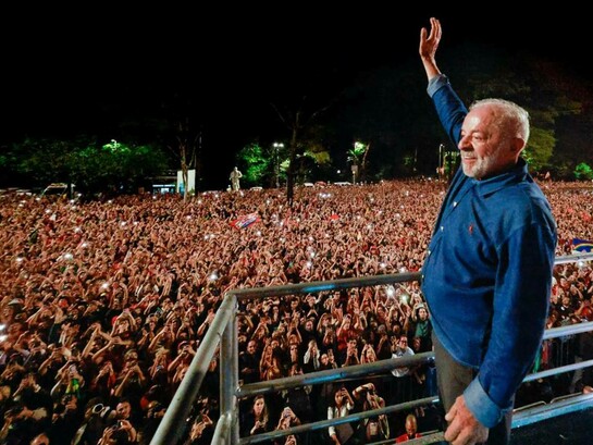 De uma plataforma, Luiz Inácio Lula da Silva ergue o braço em saudação a uma multidão que ocupa a rua e o espaço público durante a noite. Milhares de pessoas levantam as mãos e os celulares, enquanto bandeiras e manifestações de apoio compõem a cena. A imagem traduz a dimensão popular da vitória de Lula e a capacidade de mobilização que marcou sua trajetória política. Para Boaventura de Sousa Santos, porém, a vitória eleitoral não encerra a disputa em torno do futuro do Brasil: ela recoloca no centro do debate a defesa da democracia, o enfrentamento da extrema-direita e a tarefa de transformar o apoio popular em participação política efetiva