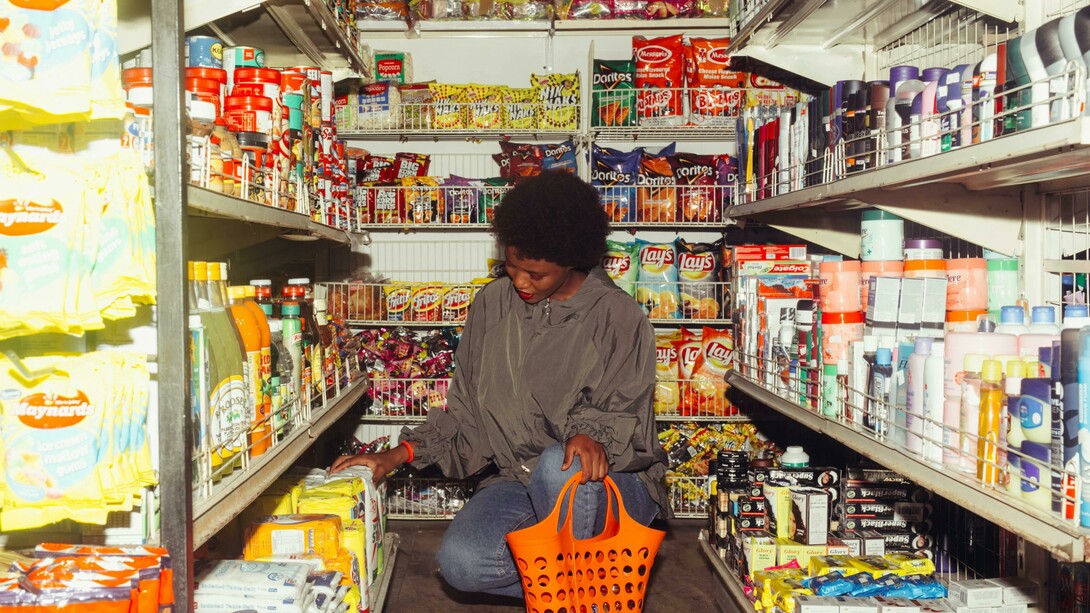An African American woman selects products while shopping in a convenience store, carrying an orange shopping basket