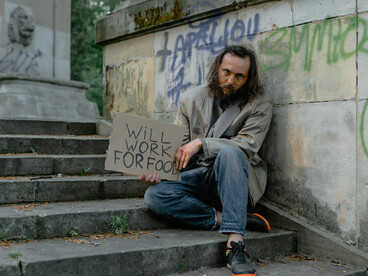 A homeless man in blue denim jeans sits quietly beside a white short coat on the sidewalk in the USA