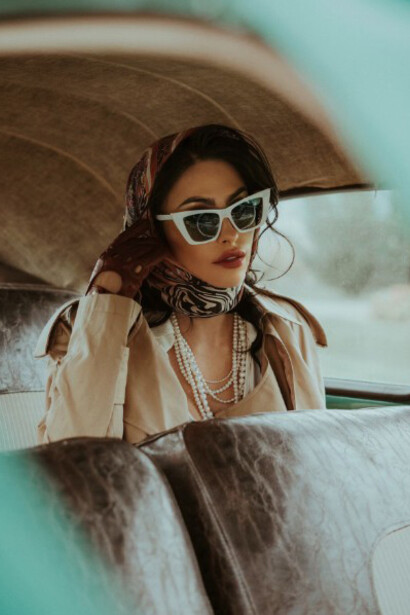Sitting in the back seat of a timeless car, a woman with sunglasses and a scarf embraces the nostalgic vibe of the moment