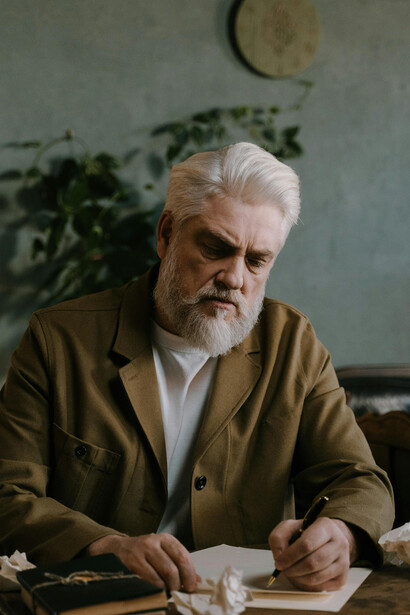 An elderly man sits thoughtfully at a table, holding a pen as he reflects and writes