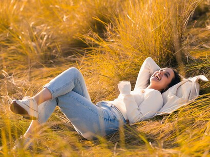 Deitada entre a vegetação, a mulher sorri enquanto desfruta da tranquilidade da paisagem natural. Na espiritualidade associada a Gaia, estar presente na natureza é também reconectar-se consigo mesmo