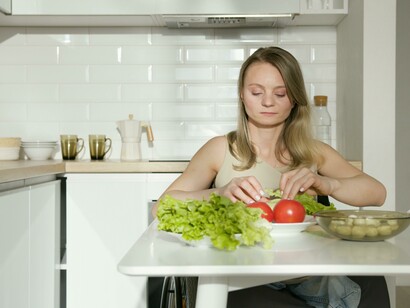 A woman in a beige tank top sitting in the kitchen beside a table of vegetables
