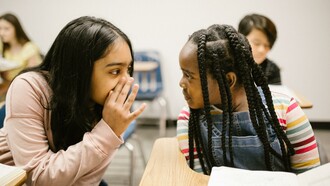 Two girls whispering during a lesson, inside a classroom, at school
