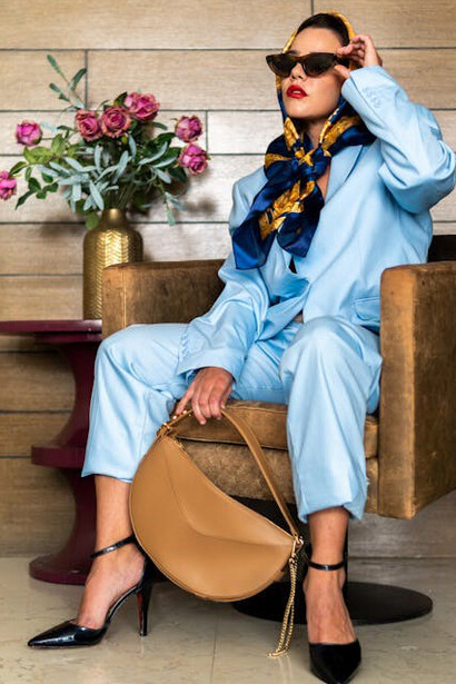 Poised and stylish, a woman sits effortlessly in a light blue suit, adorned with a matching headscarf, trendy sunglasses, and elegant black heels
