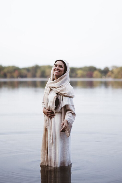 A woman stands in the water, wearing a biblical robe and smiling