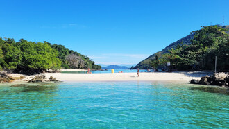 A praia aparece entre o mar, a vegetação e as montanhas ao fundo, formando uma paisagem de águas tranquilas e natureza exuberante. Em Ilha Grande, o afastamento do ritmo urbano abre espaço para desacelerar e reencontrar outros modos de estar no mundo