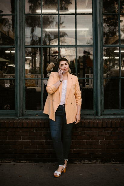 A woman dressed in a beige coat and black pants stands outside a building at night, engaged in a phone conversation that reflects a long-distance relationship