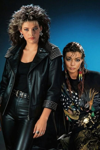 Two women showcase the bold style of the 1980s, featuring voluminous hair, leather jackets, and the decade’s signature confidence