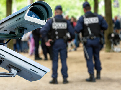 A surveillance camera monitors a public area while police officers patrol in the background