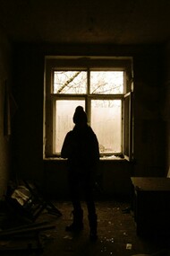 A solitary figure stands silhouetted by a window in an abandoned house, surrounded by fading light and quiet decay