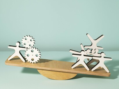 A seesaw is the central element of this image, with gears and human-shaped figures balancing on either side, suggesting a moment of equilibrium, but also hinting at the potential for change