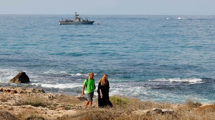 People walking along the beach as a navy vessel patrols the Mediterranean waters off Rosh Hanikra, known in Lebanon as Ras al-Naqura, on the Israeli side of the border between the two countries, on October 7, 2022