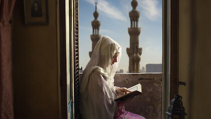 A woman in Egypt practices her worship rituals inside her home during Ramadan, with the two prominent minarets in the distance belonging to the Al-Rifa'i Mosque in Cairo, Egypt