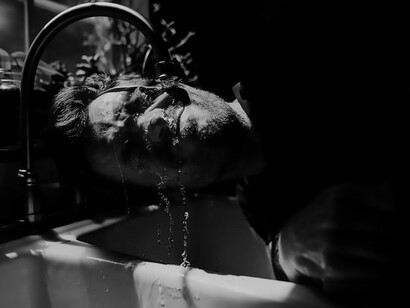In a striking visual tableau, a man submerges beneath the streaming water from a tap, creating a powerful image that speaks to the immersive nature of daily rituals and the elemental essence of life