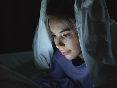 Sob o cobertor, a mulher utiliza um tablet durante a noite. O quarto, tradicionalmente associado ao repouso, também se transforma em espaço de consumo de conteúdo e prolongamento do tempo diante das telas