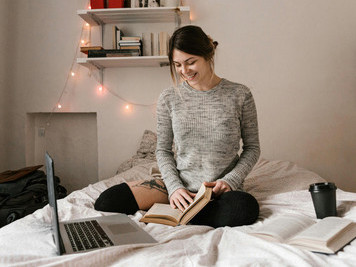 A woman in a gray sweater sits on her bed with a laptop and several books in a calm, minimalist room