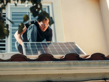 A man installs a solar panel while standing on the roof of a house