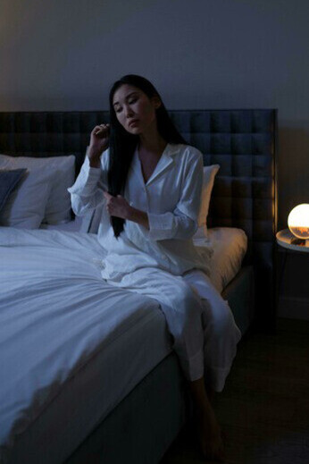 A woman in white sleepwear sits on the edge of a bed, caught in a sleepless night