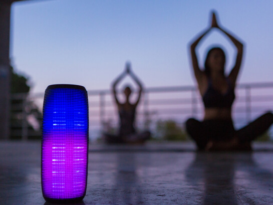 A colorful backlit wireless speaker in the foreground, as a group of friends practices yoga in the background