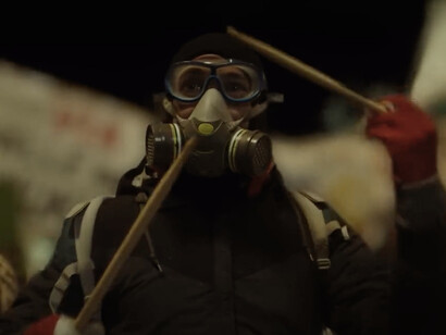 In the midst of the protest, the ominous figure of a gas mask-clad demonstrator stands out, a silent protester ready to face the inevitable confrontation with the riot police