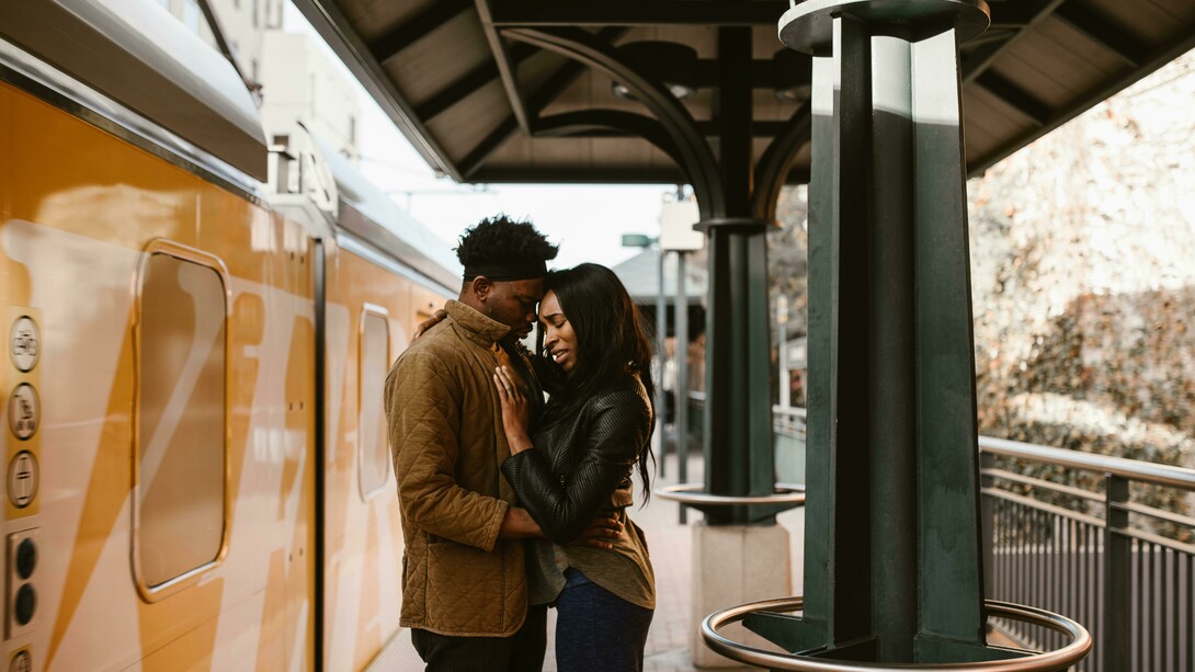 A couple hugs beside an orange train, sharing an emotional moment in a long-distance relationship