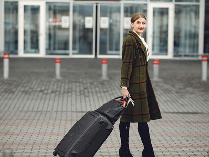 A stylish woman walks past a contemporary building, carrying luggage, a passport, and a boarding ticket