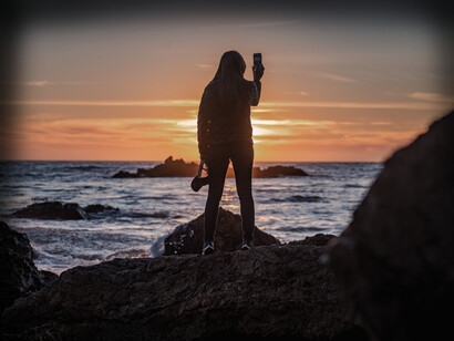 The sun is setting on the sea as a woman takes a photo