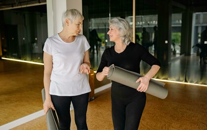 Two senior women chatting and smiling while holding yoga mats inside a modern fitness studio