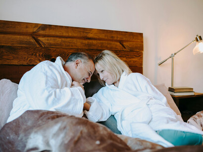 An elderly couple in white robes lying on a bed, sharing a joyful kiss
