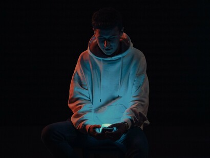 Um jovem vestido com um moletom aparece em um ambiente escuro, com o rosto iluminado pela luz do celular que segura nas mãos. O contraste entre a tela luminosa e a escuridão ao redor reforça a sensação de isolamento. A cena remete ao tédio digital, que pode nos levar a permanecer diante do aparelho mesmo quando já não encontramos nele algo que realmente desperte nosso interesse