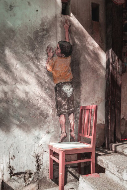 Wall art of a boy in black shorts standing on a red wooden seat in George Town, Malaysia