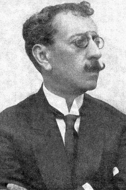 Olavo Bilac em um retrato de braços cruzados, usando terno, gravata e óculos. A imagem remete à figura de um dos principais representantes do parnasianismo brasileiro, movimento que valorizava o rigor formal e o trabalho minucioso com a linguagem, aspectos evidenciados em Profissão de fé
