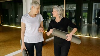 Two senior women chatting and smiling while holding yoga mats inside a modern fitness studio