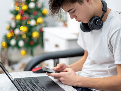 A teenage boy uses a smartphone with headphones while balancing a laptop on his knees at home, illustrating the impact of technology on children and the prevalence of digital distractions
