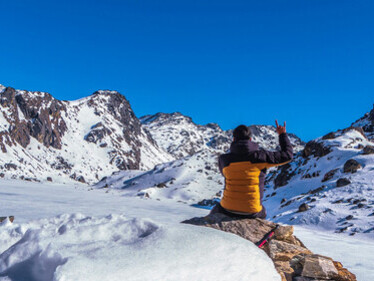 A person in a blue jacket and orange pants sitting on a snow-covered rock formation during the daytime, by the freezing Gosaikunda Lake, Nepal