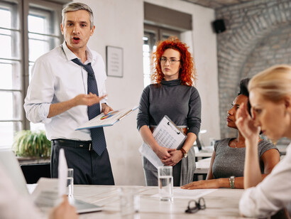 Un uomo sta animatamente presentando dei documenti o delle idee a due colleghi seduti, durante una riunione o una discussione di progetto
