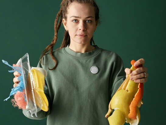 Uma mulher segura resíduos plásticos em uma mão e resíduos orgânicos na outra, evidenciando dois destinos possíveis para os materiais descartados. Entre o passivo ambiental e a matéria-prima do futuro, a escolha define o modelo de desenvolvimento. A imagem simboliza o contraste entre a linearidade do descarte e o potencial regenerativo da biomassa. A transição para uma economia circular começa na forma como enxergamos o lixo