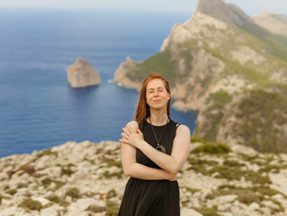 A red-haired woman stands with her eyes closed on a rock, gazing at the breathtaking mountain views of the Formentor Peninsula in Mallorca
